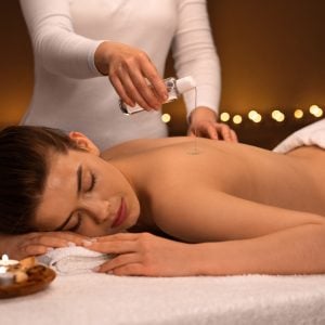 Massage therapist pouring oil on back of lady with lit candles in background.