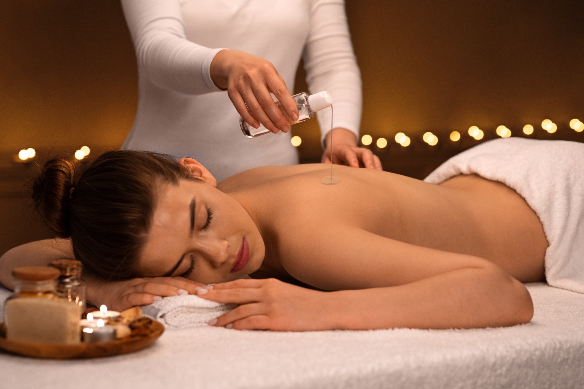 Massage therapist pouring oil on back of lady with lit candles in background.