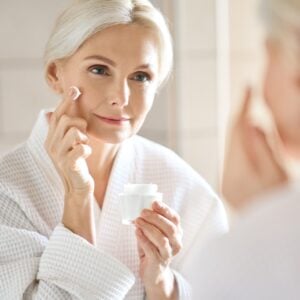 Middle age woman in white bath robe applying face skin care cream treatment while looking in a mirror.