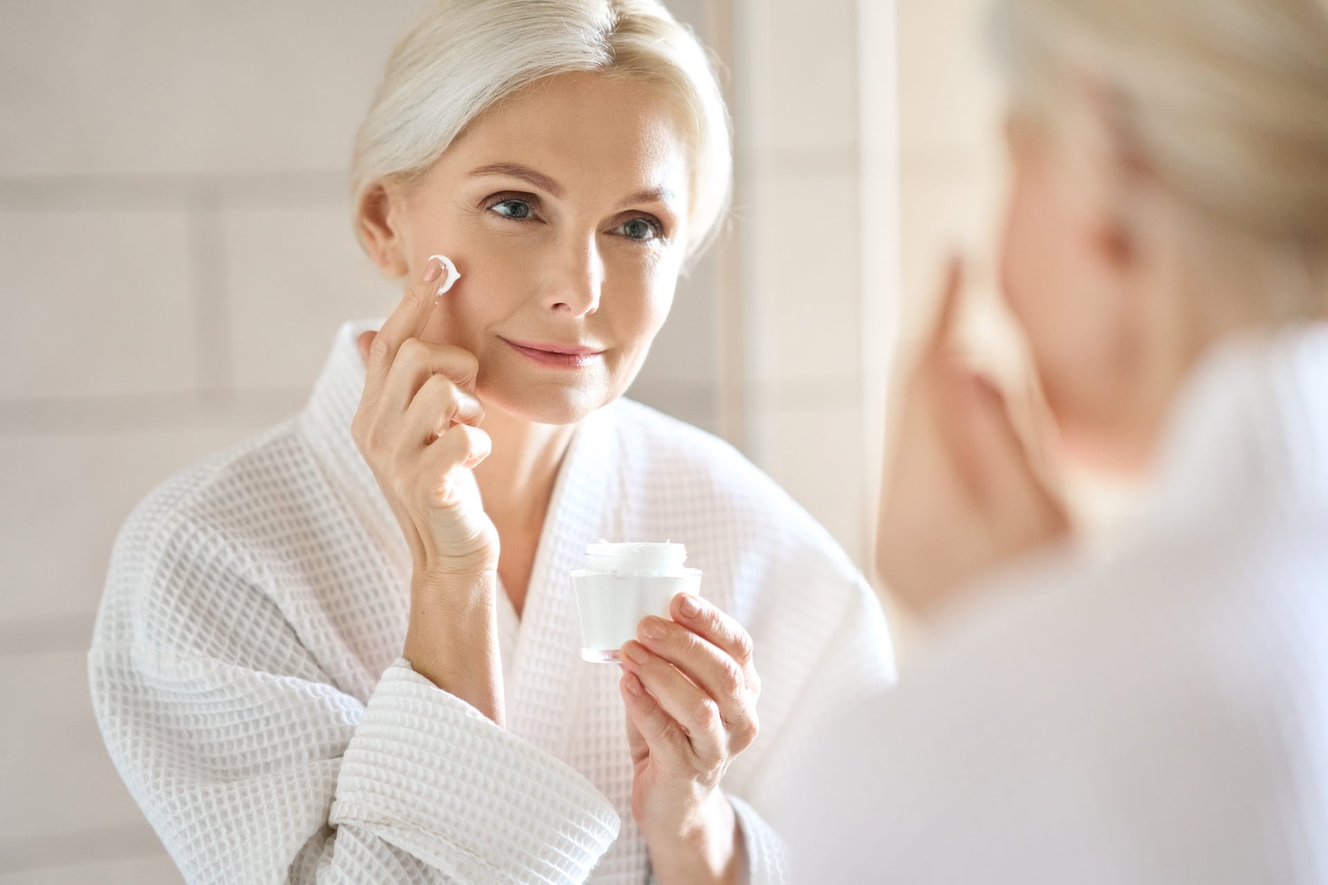 Middle age woman in white bath robe applying face skin care cream treatment while looking in a mirror.