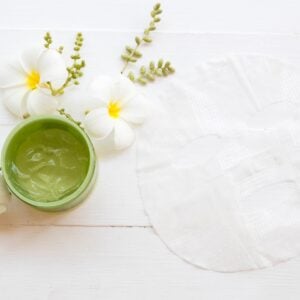 Green facial mask in small jar with white flowers in the background.