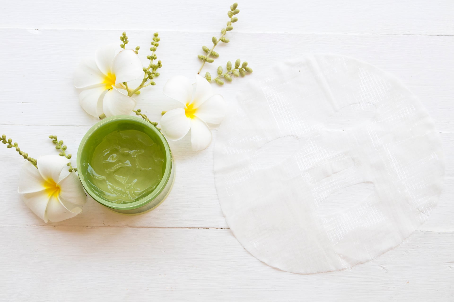 Green facial mask in small jar with white flowers in the background.
